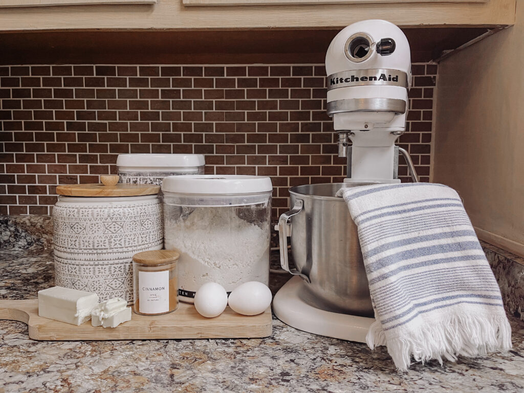 white Kitchen Aid Mixer with tea towel next to cutting board and sugar canisters, two eggs, butter, and jar of cinnamon-2