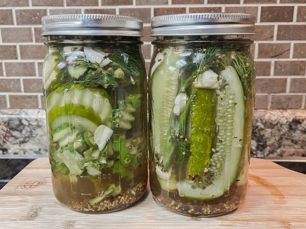 Two jars of freshly made pickles on wooden cutting board against brown subway tile background